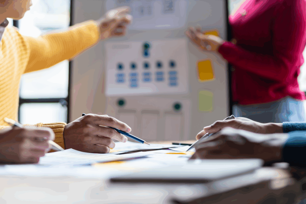 Pessoas reunidas em uma mesa de trabalho analisam documentos e fazem anotações enquanto outra pessoa aponta para um quadro com fluxogramas e post-its coloridos ao fundo, durante um processo de diagnóstico no plano de governo.