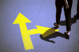 Pessoa caminha ao lado de uma seta amarela no asfalto que se divide em duas direções, representando escolhas no contexto dos gatilhos mentais na construção do plano de governo.