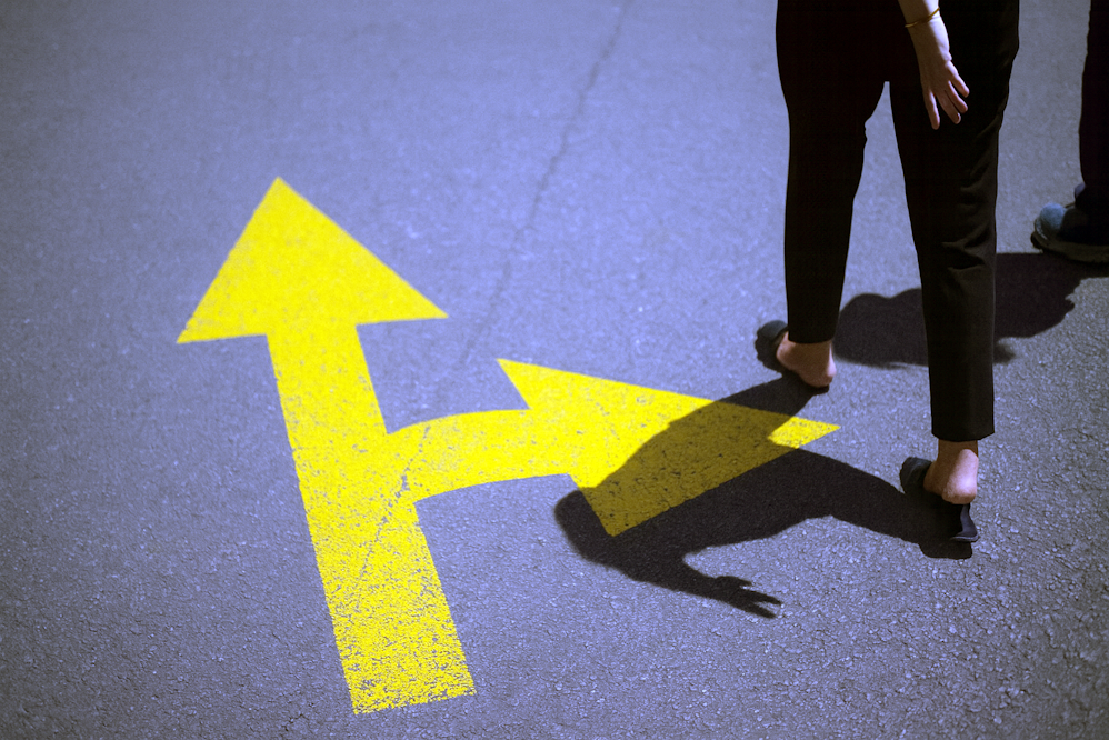 Pessoa caminha ao lado de uma seta amarela no asfalto que se divide em duas direções, representando escolhas no contexto dos gatilhos mentais na construção do plano de governo.