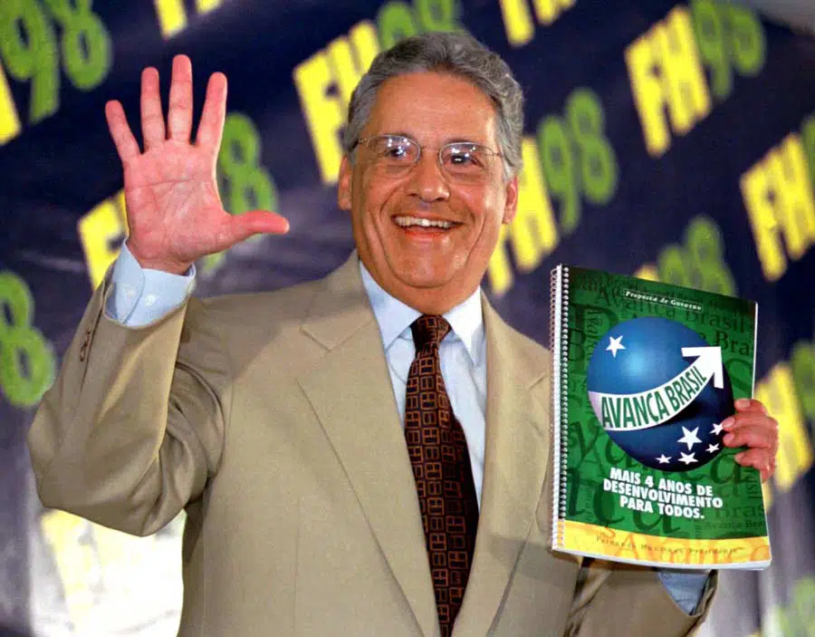 Fernando Henrique Cardoso sorri, levanta a mão com os cinco dedos abertos e segura o caderno do plano de governo FHC 1998, com a marca “Avança Brasil” em destaque ao fundo.