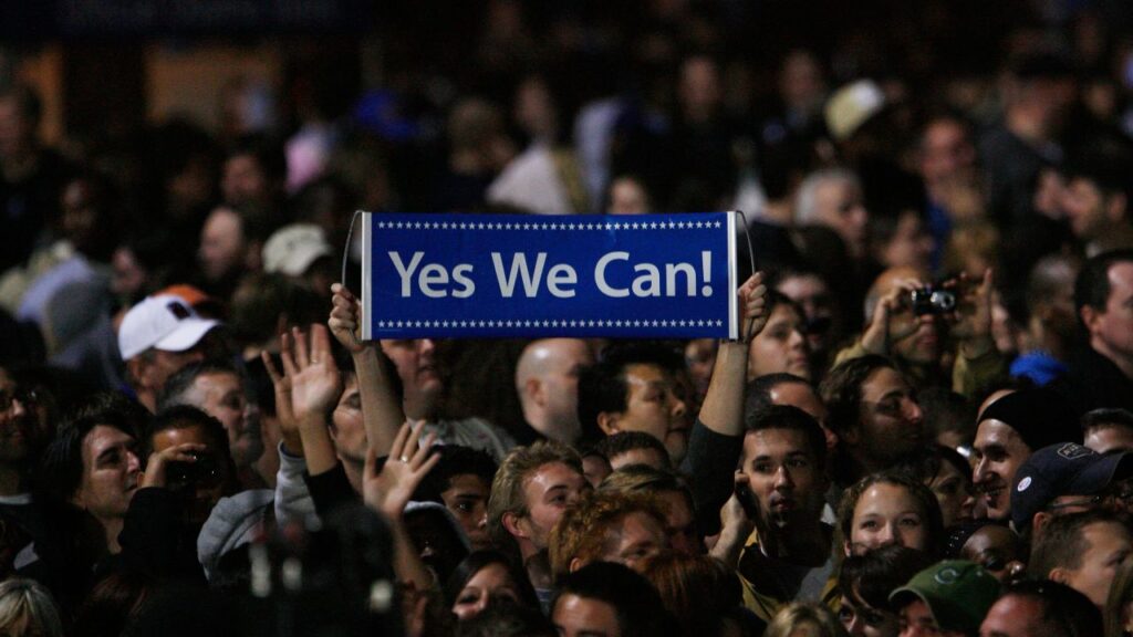 Multidão em comício de Barack Obama segurando uma placa com os dizeres "Yes We Can!" (Sim, nós podemos), slogan central do plano de governo Obama