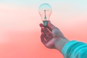 Pessoa segura uma lâmpada acesa diante de um céu com tons rosa e azul, representando o surgimento de ideias e soluções criativas — imagem simbólica para o conceito de design thinking no plano de governo.