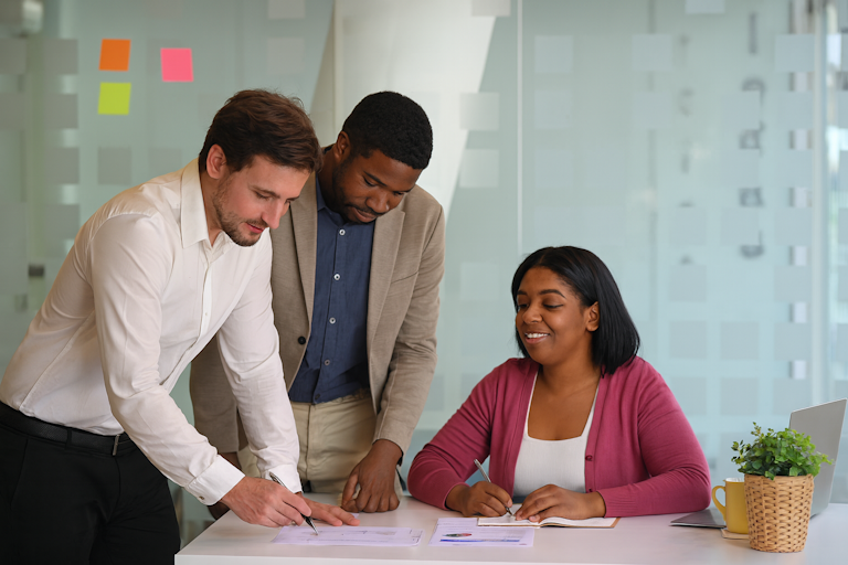 Três assessores brasileiros, dois homens em pé e uma mulher negra sentada à mesa, analisam documentos e gráficos sobre um plano de governo para candidatos sem mandato no Executivo em uma sala de reunião moderna, com notebook, caneca e post-its coloridos ao fundo.