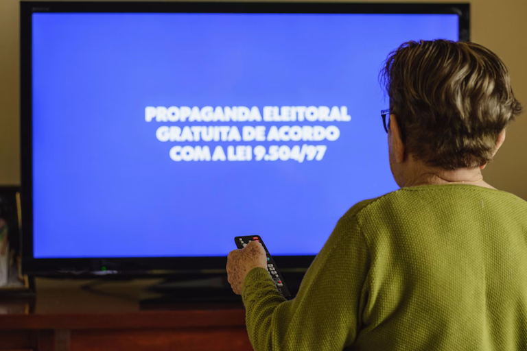 Eleitora assiste à propaganda eleitoral gratuita na televisão, com a mensagem sobre a Lei nº 9.504/1997 exibida na tela durante o horário eleitoral.